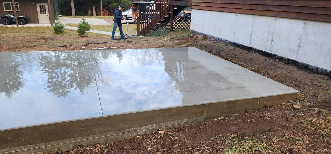 The image showcases a recently completed concrete slab reflecting trees and sky on its water-covered surface. A person, standing on one edge, appears to be washing or treating the slab. In the background, there's a green ATV and a house with a white porch.