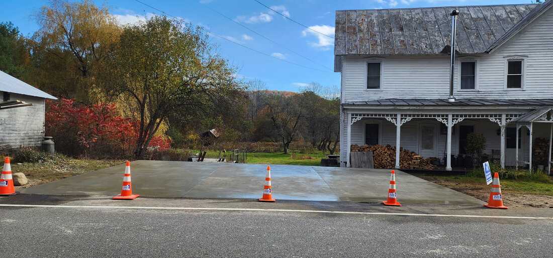 A large, finished concrete slab extending from a roadway with traffic cones outlining its border. The area is sunny with clear skies, and a white house with a covered porch and a woodpile is visible in the background, alongside autumnal trees.